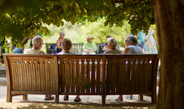 A group of older people on a bench in full sun surrounded by trees