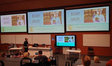 A group of people look at a screen with 'botany careers' on it. A presenter is giving at talk in front of it.